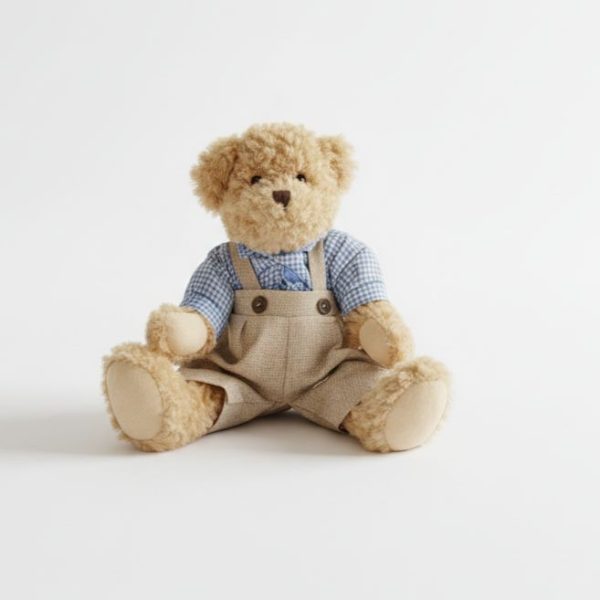 Oso de peluche con camisa celeste y jardineras beige sobre fondo blanco